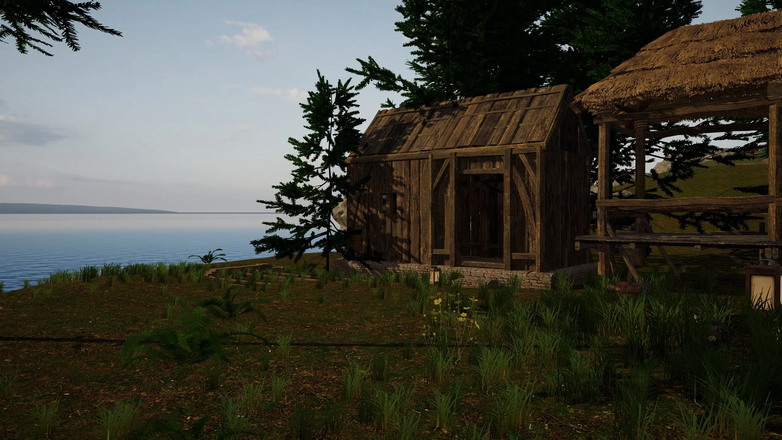 The finished Level 2 timber house on its stone footing, next to the thatched starter shelter, with the sea beyond.